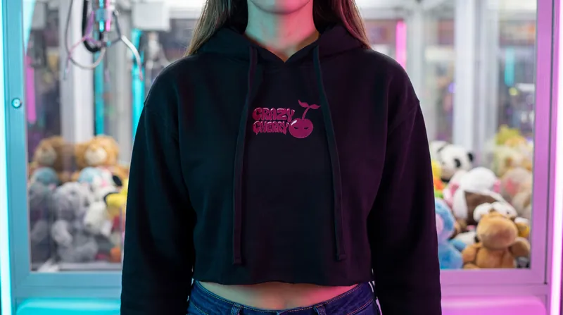 A black cropped hoodie with a small chest logo, worn in front of a claw machine.