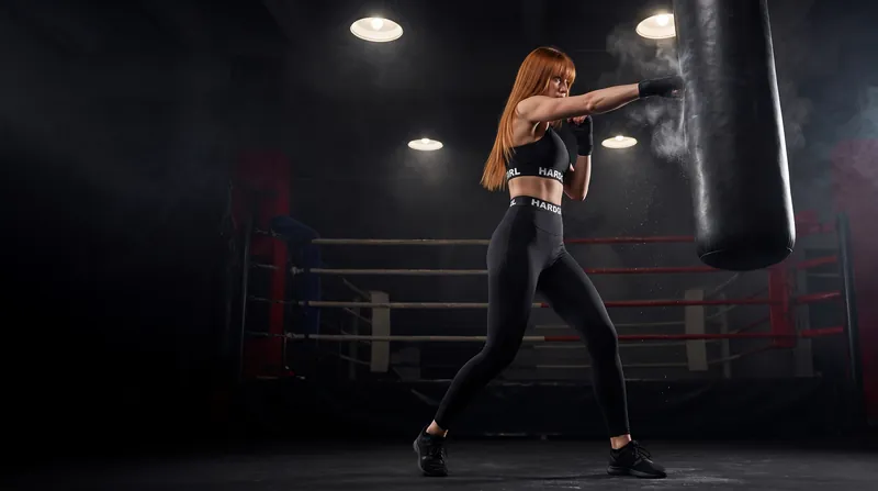 A red-haired woman in black training kit throws a punch at a heavy bag beside a boxing ring.